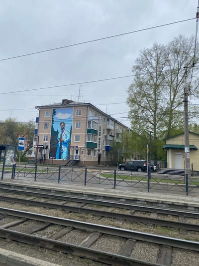 The image shows an exterior view in Usolye-Sibirskoye, Russia. An apartment building with a large mural depicting a man in a medical coat is visible. There's an adjacent trolleybus stop with signage. A black car is parked along the street. Trees are present, and trolley tracks run in the foreground. The sky appears overcast.