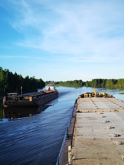 A tranquil, wide-angle photograph captures a peaceful waterway scene near Annensky Most, Russia. The perspective is from the deck of a large, flat-topped cargo barge, partially visible along the right side of the frame, featuring a blue railing and numerous circular hatches on its concrete-like surface.
In the mid-ground, a second, complete cargo vessel consisting of a dark-hulled barge being pushed by a white and red-trimmed tugboat with a prominent cabin, navigates away from the viewer. This vessel creates a distinct V-shaped wake that ripples across the calm, dark blue water. Both banks of the river are densely lined with lush, vibrant green forests, primarily composed of tall, slender trees, which are partially reflected in the water's surface.
The sky above is a bright, clear blue with sparse, wispy white clouds. The natural lighting suggests either early morning or late afternoon, as a warm, low-angle sun illuminates the trees and the visible vessel, casting subtle shadows and highlighting the textures of the water. No people are clearly discernible on either vessel or along the riverbanks. There is no legible text visible anywhere in the image. The overall atmosphere conveys serene navigation through a natural, wooded landscape.