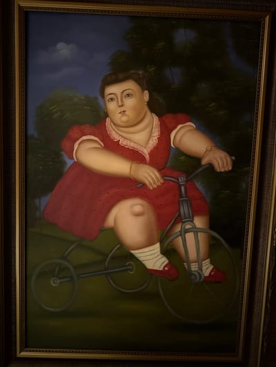 A full-body oil painting depicts a woman with an ample physique riding a bicycle. She is wearing a red dress adorned with small dark polka dots and a white ruffled collar. Her legs are clad in white socks with blue stripes and she wears red shoes. The woman has fair skin, dark hair pulled back with a red ribbon, and her face is turned slightly to the left. She is wearing gold bracelets on both wrists and green earrings. The bicycle is dark-colored with thin wheels. The background features dark green trees against a twilight sky with faint white clouds. The painting is framed by an ornate, textured frame. The scene is not located in the city of Heiligenrode, Germany.