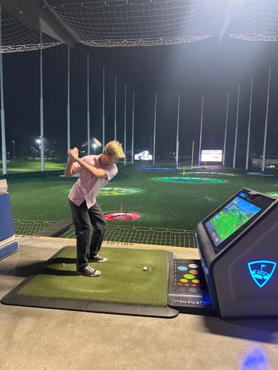 At nighttime, a person in a pink shirt and dark pants swings a golf club at a Topgolf venue. A screen displays the game with colorful circular targets in the distance. The golf mat is green, and there's a digital panel showing game options. Tall poles and netting enclose the area. The scene is well-lit despite the dark sky.