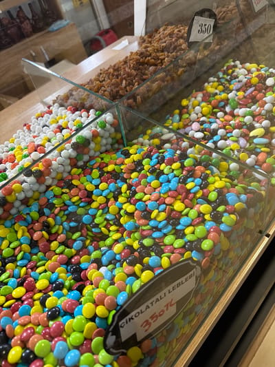 The image shows a close-up of a display of candies in what appears to be a market or shop. Two distinct types of candies are visible. In the foreground, a large bin is filled with small, colorful, coated candies in shades of red, blue, yellow, green, pink, white, and black. These candies are round or slightly oval. Behind this bin, and separated by glass, is another bin filled with white and light-colored candies, also appearing to be coated, possibly dragees. Further back and to the right, a bin is filled with dried fruit, likely raisins, which are a light brown color. A price tag is visible on the display of the dried fruit, showing the number "350". Another price tag is visible on the display of the colorful candies, which reads "ÇİKOLATALI LEBLEBİ" (chocolate coated roasted chickpeas) and "350₺". The lighting suggests an indoor setting with artificial lights. The context provided suggests this scene is located in Acıpayam, Turkey, likely within a shop selling confectionery and dried goods. There are no people or specific actions depicted, only the static display of products.