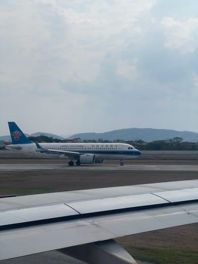 The image captures an airport scene from the perspective of an aircraft's wing, which dominates the right side of the frame. On a parallel runway or taxiway, a China Southern Airlines airplane is visible, heading towards the right. The airplane is white with blue and red accents; "CHINA SOUTHERN" and its Chinese equivalent, "中国南方航空", are printed on its fuselage. The tail fin features the airline's distinctive red Kapok flower logo on a blue background. The text "AIRBUS A320" can also be seen on the main wing of the China Southern aircraft, indicating its model.
In the background, a range of mountains rises under a vast, partly cloudy or hazy sky. The foreground and midground feature the asphalt of the runway/taxiway and some sparse grassy areas. The scene takes place during daylight hours, characterized by soft, natural illumination, and suggests an overcast or slightly hazy weather condition. No people are visible. The overall setting is an airport in a mountainous region, aligning with the provided location context of Ban Rong Ruea Kham, Thailand.