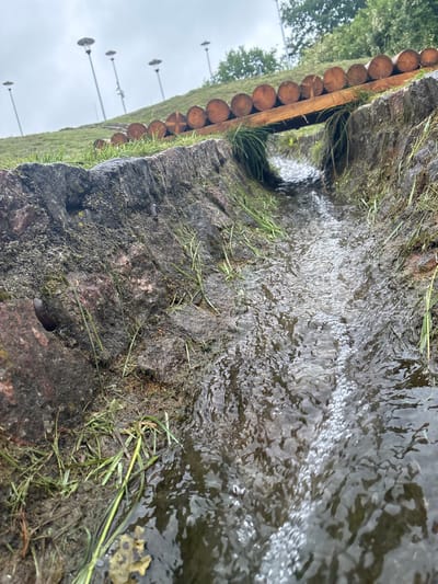 A close-up, low-angle view captures a small, rocky water channel with a wooden bridge constructed from logs overhead. The channel is filled with flowing water, creating ripples and aeration. Lush green grass grows along the edges of the channel and the surrounding earthy bank. In the background, a grassy slope is dotted with modern-style street lamps. The scene appears to be an outdoor public space, possibly a park or a scenic walkway in Sabile, Latvia, suggesting a man-made water feature integrated into the natural landscape.