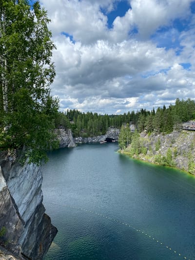 The image depicts the Marble Canyon, a flooded former quarry located in Ruskeala, Russia. The canyon features steep, sheer marble cliffs plunging into deep, clear turquoise water. Dense evergreen forests line the upper rim of the canyon, with some deciduous trees, including birches with their white bark, visible on the left foreground. The sky is partly cloudy with dramatic grey and white cumulus clouds interspersed with patches of blue.
In the water, several small boats are visible, some appearing to be navigating towards or within a cave opening in the far left cliff face. A line of yellow buoys stretches across the water, suggesting a designated swimming or boating area. On the right side of the canyon, a wooden boardwalk is visible, elevated along the cliff, with people walking and observing the scenery. The overall impression is of a natural landscape transformed by quarrying, now a popular tourist destination. The lighting suggests it is daytime, likely late morning or afternoon, under a dynamic sky.