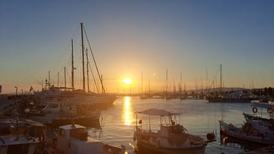 A wide shot captures a serene marina scene at either sunrise or sunset in Alimos, Greece, characterized by a vibrant golden glow across the sky and water. The sun, a brilliant orb with some lens flare, is low on the horizon, positioned slightly to the right of the image center, casting a long, shimmering path of light across the calm water surface towards the viewer.
The sky transitions from a bright, warm orange and yellow near the horizon, gradually fading into a clear, soft blue overhead. Numerous boats of various sizes fill the marina, their silhouettes, masts, and rigging creating an intricate pattern against the bright sky. In the foreground, several smaller motorboats or fishing boats are docked, some with covers or awnings. Details like ropes, bumpers, and a white life ring are visible on these boats.
In the midground and background, a multitude of sailboats with tall masts and extensive rigging, alongside larger motor yachts, are moored. One prominent, large sailboat with multiple masts stands out on the left side of the frame, with a flag, possibly Greek, visible on its stern. The distant shoreline on the right features more boats and what appear to be low-lying hills or landmasses, also silhouetted. A dock or pier with light posts and indistinct structures is visible on the far left, bordering the moored vessels. The overall atmosphere is peaceful and tranquil, with no discernible human activity. No visible text can be clearly identified in the image.