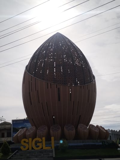 The image depicts a large, oval-shaped monument or structure in Lampeudeu Baroh, Indonesia. The monument is primarily brown and features a latticework design at the top and vertical slats on the lower portion. The base consists of multiple smaller, similarly designed oval shapes. The word "SIGLI" is spelled out in yellow letters in front of the monument. Overhead power lines crisscross the sky, which is overcast. In the background, there are buildings and a billboard with text and an image. A small amount of greenery is visible at the base of the monument.
