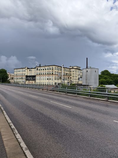 The image depicts the former Cloetta factory in Ljungsbro, Sweden, on a cloudy day. A paved road with white lane markings occupies the foreground, with a concrete sidewalk on the left. A metal guardrail separates the road from the factory complex. The factory building is a large, light-colored structure with multiple stories and numerous windows. To the right of the main building is a tall, cylindrical structure with a smokestack. Trees and greenery are visible behind the factory. The sky is filled with dark, ominous clouds, suggesting impending rain or a storm. No people are visible in the image.