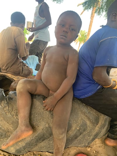 The image, taken in Gobbiya, Nigeria, depicts a close-up view of a young child, appearing to be a boy, with dark skin and short, dark hair. He is lying down, with his torso and legs exposed and covered in a thin layer of dust or dirt. His eyes are open and looking towards the viewer. The child is positioned on what appears to be a large, dark, rough object, possibly a piece of tarpaulin or a tire.
In the background, other individuals are visible. To the left, a person wearing a blue shirt is partially visible, with their arm adorned with several bright orange bracelets. Another person, wearing a light blue tank top and brown pants, is also in the background, holding a metal cup. Further back, another person is seated, wearing a brown garment. The setting appears to be outdoors, with hints of palm trees and bright, possibly sunny, daylight. The ground is dusty and uneven, with scattered dry leaves. There is no visible text in the image. The overall impression is one of a casual, outdoor gathering, possibly in a rural or informal settlement.