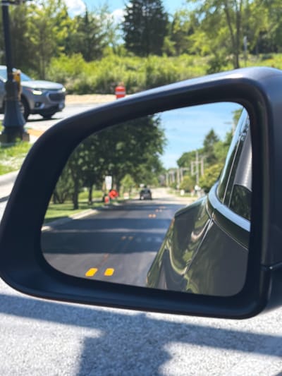 A side-view mirror of a vehicle is shown, reflecting a street scene in Buffalo Grove, Illinois. The mirror reflects a road with cars, trees, and utility poles. Two orange traffic cones are visible in the distance. The vehicle's blind-spot monitoring system indicator lights are visible in the lower part of the mirror. The background shows a sunny day with green trees and a blue sky.