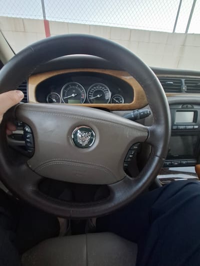 A close-up shot captures the steering wheel and dashboard of a Jaguar car. The steering wheel is dark brown leather, with a light brown leather center pad emblazoned with the Jaguar emblem. The dashboard features analog gauges displaying speed and other vehicle information, surrounded by wood trim. A control panel with various buttons is visible above the steering wheel. A person wearing a navy blue shirt is partially visible in the right side of the image. The environment appears to be inside a car, and the location is unknown.
