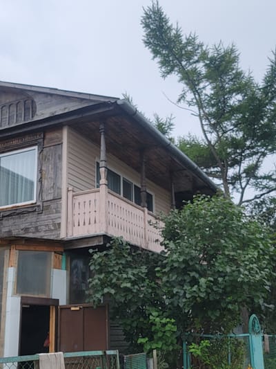 The image shows a weathered, wooden house with a light-colored siding upper floor and a dark-colored porch roof supported by wooden posts. A light pink balcony railing with decorative cutouts is visible on the upper floor. A large tree with green foliage grows in front of and to the right of the house. The sky is overcast and gray. The lower part of the image shows a portion of a turquoise metal fence, some overgrown vegetation, and what appears to be an open doorway leading into a dark interior. A piece of beige fabric hangs from the fence. The image is taken from a slightly low angle, looking up towards the house and tree. This scene is located in Timiryazevskiy, Russia.