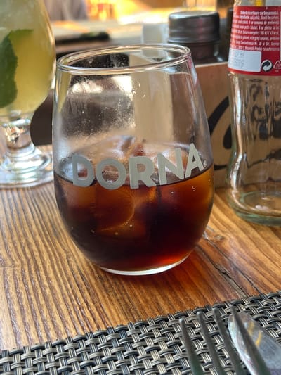 A close-up shot focuses on a glass of dark soda with ice, resting on a wooden table. The glass is partially filled with the beverage, and ice cubes are visible within. The word "DORNA" is emblazoned in silver lettering on the side of the glass. To the left of the glass, another glass containing a light green beverage with a lime slice is partially visible. In the background, to the right, a clear glass bottle, possibly for soda or water, stands empty. The table surface is a warm-toned wood with a prominent grain pattern, and a woven placemat is visible in the foreground. The lighting suggests an outdoor or well-lit indoor setting. The image does not provide any specific visual cues to determine the city of Otopeni, Romania, nor does it depict any people or actions.