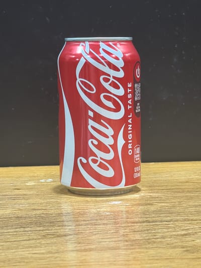 A red Coca-Cola can is laying on its side on a surface that is half black and half light brown wood grain. The can's label is visible and reads "Coca-Cola ORIGINAL TASTE 140 CALORIES PER CAN 12 FL OZ (355ml)". The image does not provide information on the location in Niles, United States of America or any information about people, actions or interactions.