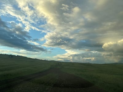 An overcast day in Elimbetovo, Russia, where a dirt road winds through rolling green hills. Power poles are sparsely scattered across the landscape. The sky is filled with dramatic, voluminous clouds in varying shades of white, gray, and blue, with hints of sunlight breaking through in some areas. The overall impression is one of vast, open space under a dynamic sky.