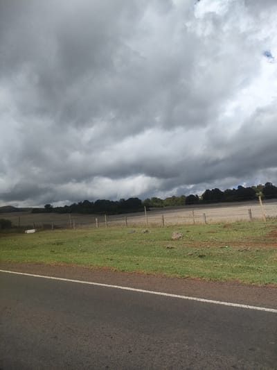 The image depicts a rural landscape in Kisima ward, Kenya, under a heavy, overcast sky. A paved road with a solid white line marking the edge runs along the bottom of the frame. To the right of the road is a grassy verge that transitions into a field enclosed by widely spaced wooden posts, presumably a fence. The field is mostly brown and appears to be recently cultivated or fallow. A line of trees is visible in the background, marking the horizon. There are no people, animals, or vehicles visible in the scene. A single white object is visible towards the left in the field.