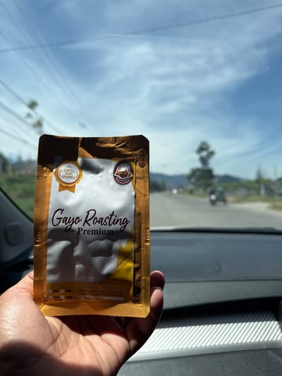 A person's hand holds a golden-brown foil package of "Gayo Roasting - Premium" coffee in front of a blurred background of a road with distant mountains under a bright, cloudy sky. The package has circular seals and text indicating "100% KOPI ARABIKA." The scene appears to be viewed from inside a vehicle, suggested by the dashboard and car interior in the foreground and lower portion of the image. The location is identified as Lemahburbana, Indonesia.