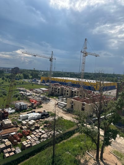 The image shows a construction site in Rubtsovo, Russia, under a cloudy sky. In the foreground, a tall, yellow tower crane is positioned on the right, with its boom extending horizontally. To its left, another, older, red tower crane is visible, with its boom tilted upwards. In the middle ground, the partially constructed brick buildings of an apartment complex are seen, with multiple floors showing exposed brickwork and unfinished windows. Adjacent to the apartment buildings is a large, bright yellow and blue commercial building. The ground around the construction is a mix of dirt and sparse grass. Piles of construction materials, including stacks of concrete slabs, bricks, and wooden pallets, are scattered across the site. In the background, more apartment buildings and a wooded area are visible, suggesting a semi-urban or suburban setting.