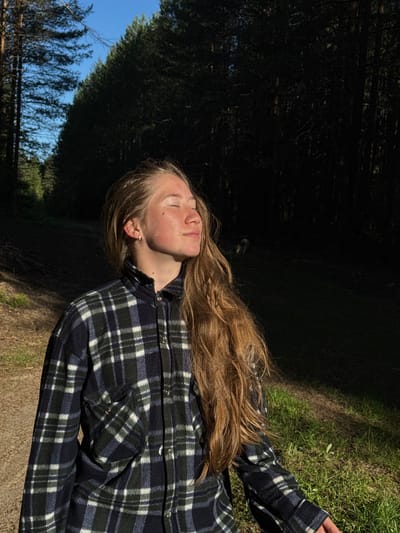 The image, taken in Ust'-Silayka, Russia, shows a young adult female standing in a forested outdoor setting. Her head is tilted slightly upward and to the right, with her eyes closed, as direct sunlight illuminates the right side of her face and her long, wavy, light brown hair. She exhibits freckles on her face and is wearing a dark blue, dark green, and white plaid long-sleeved shirt, which appears to be made of a thick, possibly flannel or fleece, material. The shirt has a single buttoned chest pocket on the left side. A small silver hoop earring is visible in her left ear. She stands on a dirt path with scattered dried leaves, adjacent to a patch of vibrant green grass to her right that is also illuminated by sunlight. The background features a dense evergreen forest, predominantly in deep shadow, with a narrow strip of blue sky visible above the treeline on the left. A faint, blurry animal, possibly a dog, is discernible in the deep shadows of the forest in the far background on the right.