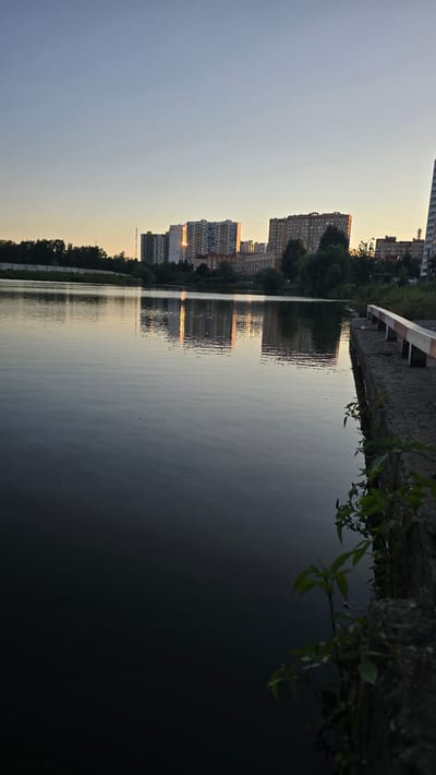 The image shows a serene, waterfront scene at dusk in Ashcherino, Russia. The main subjects are a row of tall, residential apartment buildings lining the opposite bank of a calm body of water. The buildings are of varying heights and architectural styles, with many windows visible. Their reflections are clearly mirrored in the still water, creating a symmetrical and tranquil effect.
The activity taking place is a peaceful observation of the sunset. The time of day is evident from the soft, warm glow of the setting sun reflecting off the buildings and water, casting long shadows and illuminating the sky with hues of orange and yellow against a fading blue. The weather appears clear and calm, as indicated by the undisturbed surface of the water.
Notable details include the presence of trees and foliage along the embankment in the foreground, with some branches and leaves hanging over the water. The embankment itself is composed of a gravelly path and a white-and-red striped barrier. The overall mood conveyed is one of quietude and natural beauty, with the urban landscape softened by the twilight and the reflective qualities of the water. There is no visible text in the image.