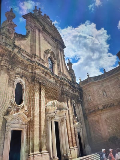 The image shows a wide-angle view of an ornate Baroque building with a blue sky and white clouds above. The building's facade is made of light-colored stone and features intricate carvings, arched windows, and several statues in niches. The architectural style suggests a historic church or public building. The perspective is from below, looking up at the building. In the lower right corner, three individuals are visible, two wearing hats and looking towards the building, suggesting they are tourists or visitors. The scene is outdoors, with the building appearing to be situated in a public space. There is no visual information in the image that would place this scene in Zrenjanin, Serbia.