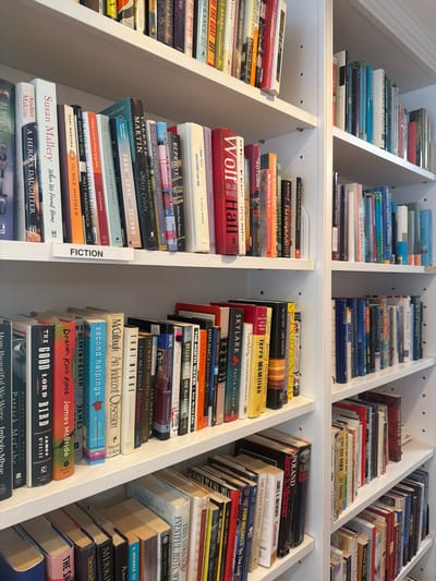 The image shows multiple white bookshelves filled with books. The bookshelves are arranged in a room with white walls and trim. A white label with the word "FICTION" is visible on the middle shelf. The books are of various sizes, colors, and thicknesses, with some showing wear and tear. The overall impression is of a well-stocked personal library or bookstore. No people are visible in the image. The location is specified as the Village of East Hampton, United States of America.