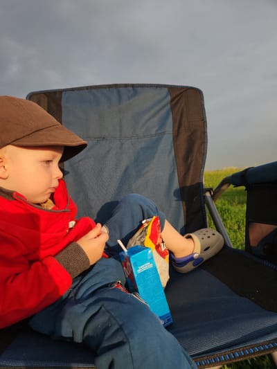 A young child wearing a brown cap, a red hooded jacket, and blue pants sits in a blue and black camping chair. The child is holding a juice box and a snack. The child's right foot is visible, wearing a beige Croc-style shoe with blue accents. The background shows a grassy field under a cloudy sky. The image was taken in Atameken, Kazakhstan.