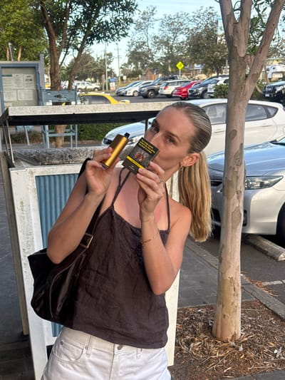 A blonde woman in a brown camisole and white jeans stands sideways, holding a yellow vape and a pack of cigarettes. She gazes at the camera. A brown purse hangs from her shoulder. A tree trunk lies horizontally in the upper portion of the frame. Cars are parked in the background.