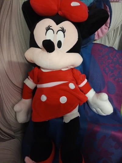 The media file shows a plush toy of Minnie Mouse. She is wearing her iconic red dress with white polka dots and a matching red bow on her head. The toy is sitting upright, positioned slightly to the left of the frame. Behind Minnie Mouse, a textured grey fabric is visible on the left, and to the right, a blue and purple patterned fabric is draped. The lighting suggests an indoor setting, possibly a home environment, with no specific time of day or weather indicated. There is no visible text in the image. The subject is solely the plush toy, and no activity or event is taking place. The background elements are indistinct and do not provide specific location cues beyond a general indoor setting.