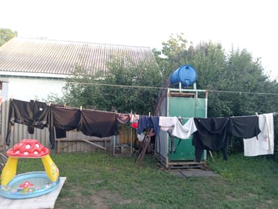 This media file depicts a backyard scene in Rakhinka, Russia, likely during the daytime with fair weather. The primary focus is a clothesline strung across the yard, laden with laundry. Various items of clothing, predominantly dark-colored, are hung to dry, secured with clothespins. These include what appear to be pants, shirts, and a towel.
In the foreground, a small, inflatable kiddie pool shaped like a mushroom is set up on a wooden platform. The pool is filled with water and contains a miniature mushroom decoration. Behind the clothesline, a weathered wooden fence encloses the yard. Further back, a residential building with a corrugated roof is visible, alongside dense green foliage from trees and bushes.
On the right side of the frame, a utilitarian structure, possibly an outdoor storage unit or shower, is present. A large blue water tank is mounted on top of this structure, suggesting a system for heating or storing water for outdoor use. The overall impression is one of domestic activity and a somewhat rustic setting. There is no visible text in the image.