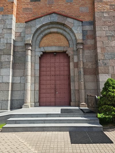 The image shows the exterior of a stone building, likely a church, with a large, reddish-brown metal double door. The door is set within a stone archway supported by two fluted stone columns. The building's facade is constructed from a combination of large, rough-hewn grey and pinkish granite blocks and red brick. A set of grey stone steps leads up to the entrance, with black rubber mats placed on the steps and at the bottom. To the right of the entrance, a small, neatly trimmed green coniferous shrub is visible, along with a black metal railing. The ground in front of the steps is paved with light-colored interlocking cobblestones. The lighting suggests it is daytime, with shadows cast on the building. This scene is the entrance to the Church of the Annunciation of the Blessed Virgin Mary in Lentvaris, Lithuania.