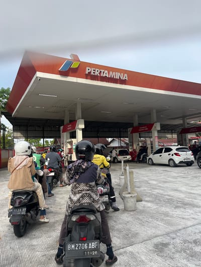 The image shows a busy gas station in Pekanbaru, Indonesia. The main subject is the Pertamina gas station itself, with its distinctive red and white branding and the Pertamina logo visible. Several people are present, mostly riding motorcycles. They appear to be waiting in line to refuel their vehicles. The scene is set outdoors, under the covered area of the gas station.
The people are dressed casually, some wearing helmets. The activity taking place is refueling at a gas station, indicated by the presence of motorcycles lined up and the overall setting. The ground is paved concrete with distinct lines.
Notable details include the time of day, which appears to be daytime, likely morning or afternoon, given the lighting. The weather seems overcast, contributing to a diffused light. There is no overt display of emotion, but the scene suggests a routine activity.
Visible text includes "PERTAMINA" prominently displayed on the building. There are also signs on the pumps, such as "PERTAMAX," "PERTAMAX TURBO," and numbers like "2" and "3" which likely indicate pump numbers or fuel types. License plates on the motorcycles are also visible, with text like "BM 2706 ABF" and "BM 4139 VY." Other signs in Indonesian are present but are difficult to read clearly.