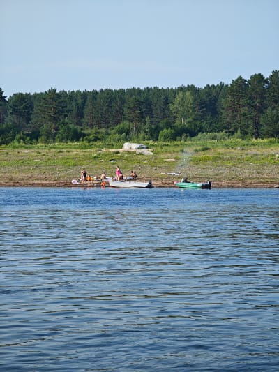 The image depicts a sunny day at a waterfront, likely in Потапово, Russia. In the foreground, a body of water with gentle ripples reflects the blue sky. The midground shows a rocky shoreline with sparse vegetation, leading up to a dense evergreen forest that forms the backdrop.
Several people are gathered on the shore and near boats. On a raft or pontoon-like structure, a group of individuals is socializing. One person in a pink swimsuit stands out. Adjacent to them, a white boat is partially visible. Further to the right, a green motorboat is docked. Smoke appears to be rising from an area near the green boat, suggesting a campfire or cooking activity.
The scene appears to be a leisure activity, possibly a picnic or a day trip to the water. The people are casually dressed, and the atmosphere seems relaxed. The time of day is likely daytime, given the bright sunlight and clear sky. The presence of boats and a forested landscape points towards a natural, possibly rural or semi-rural, setting. No visible text is present in the image.