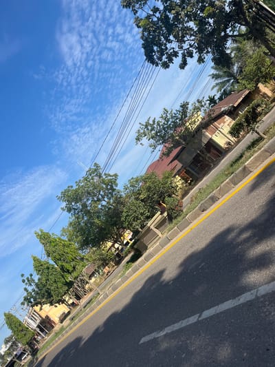 The image depicts a street scene in Tanahterban, Indonesia, on a clear day with a blue sky and scattered clouds. A paved road runs diagonally across the frame, with a yellow line marking the edge of the lane. A curb with a stone pattern separates the road from a grassy verge.
Along the right side of the street, there are several buildings with corrugated metal roofs and light-colored walls, some of which appear to be older structures. Trees and lush green foliage line the street in front of the buildings, with some taller palm trees visible further in the background. Power lines are strung across the sky, supported by poles that are not fully visible. A faint contrail from an aircraft is visible in the upper left quadrant of the sky. The perspective suggests the photo was taken from a moving vehicle or at an angle looking down at the road.
