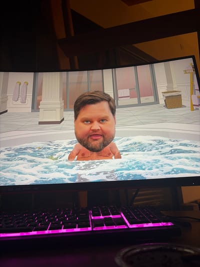 The image displays a computer monitor positioned on a wooden desk within a dimly lit indoor environment, specifically in Rosny-sous-Bois, France. The monitor, identified as an AOC brand, shows a digitally rendered scene of a male figure submerged from the chest upwards in a bubbling, light blue jacuzzi. The figure has fair skin, blue eyes, brown hair, a brown beard, and visible chest hair. His expression is neutral, with lips slightly parted. The jacuzzi water features white foam and ripples, with small green and yellow objects floating within. The background of the digital scene depicts a stylized, luxurious bathroom or spa interior with light-colored walls, white classical columns, and a tiled floor. Two light purple towels hang on golden hooks on the left wall. A dark-framed glass partition is visible in the center-right, and a woven brown laundry hamper with a white liner is positioned on the right. Below the monitor, a black mechanical keyboard with bright magenta backlighting illuminates the keys, including an "Entrée" key on the right side. A dark cable extends from the keyboard to the right. A dark, partially obscured container rests on the desk in the bottom right corner. The room's ceiling above the monitor features warm-toned wooden beams or paneling.