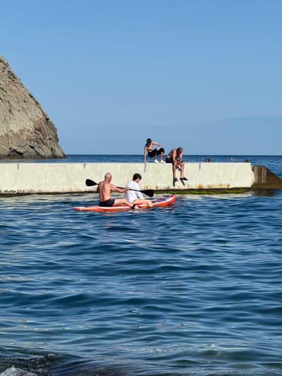 This image depicts a coastal scene in Ut'os, Ukraine. In the foreground, the blue water of the sea is visible with gentle ripples and some rocks at the very bottom. Two individuals are seated on an orange and white paddleboard in the water, with one holding a paddle. In the background, a concrete pier runs horizontally across the frame. Three other individuals are positioned on top of this pier. One is crouching, while the other two are seated, with one having their legs dangling over the edge. Further in the distance, two more people are visible in the water, appearing to be swimming. To the left of the pier, a large, rocky cliff face rises from the water. The sky above is clear and blue.
