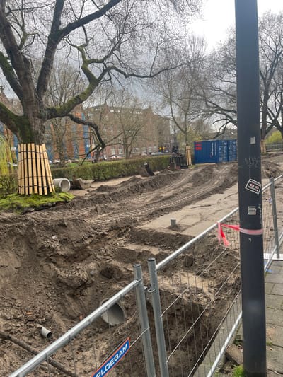 This image captures an active, large-scale urban infrastructure construction site on an overcast day in 's-Hertogenbosch, Netherlands.
In the foreground, a temporary metal mesh construction fence, bearing a "PLOEGAM" logo at its base, separates a concrete sidewalk on the right from a deep, excavated trench. A black utility pole, adorned with "SAMEN VOORUIT" and "VLOOF" stickers and wrapped with red and white striped caution tape, stands beside the fence on the right. A grey pipe section is partially visible within the trench.
The midground features an extensive area of disturbed, dark brown soil with deep tire tracks from heavy machinery. Two large concrete pipe sections, one partially visible, rest on the excavated ground. A yellow and black mini-excavator, with its bucket lowered, is situated further back. A blue shipping container, labeled "infracoep" and "infra.coep.nl," stands near the center-right, beyond the main excavation. To the left, a large, mature tree with a thick, moss-covered trunk has its base protected by vertical wooden slats secured with black straps.
In the background, a row of bare, branched trees, indicative of late winter or early spring, lines what appears to be a green space or park area. Beyond these trees, a street with parked cars is visible, followed by multi-story brick buildings featuring traditional Dutch architectural characteristics, suggesting a residential or mixed-use urban area. No people are visible in the scene.
**Location in 's-Hertogenbosch:**
The scene is consistent with ongoing infrastructure and urban renewal projects, likely along one of the city's canals, rivers (such as the Dommel), or a green corridor, where civil engineering companies like Ploegam and Infracoep are actively involved in excavation and construction work to improve waterways or public spaces.