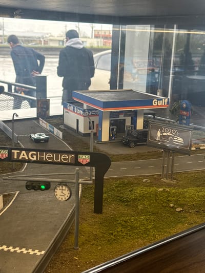 Here is a detailed description of the image:
**Scene:** The image depicts a diorama or model scene. The setting includes miniature versions of a Gulf gas station, a road with racing elements (like a starting grid), and a grassy area.
**Key Elements:**
* **Gulf Gas Station:** The miniature gas station is clearly branded with the "Gulf" logo.
* **Road and Racing Elements:** The road has white lane markers and a section with a checkered pattern resembling a starting line. There is also a TAG Heuer sign next to the road. The word "Le Mans" can be seen on a clock next to the race track.
* **Vehicle-related details:** There is a 2020's advertisement for vehicles.
**Reflection:**
* **People:** The blurred reflections of a man and possibly other figures can be seen in the glass.
* **Setting:** The location appears to be Siktarva, Georgia due to the Gulf station writing.