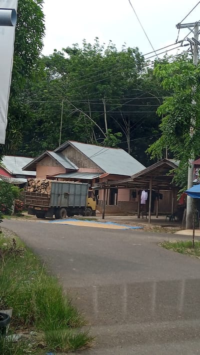 A residential area in Kebonbaru, Indonesia, features a single-story house with a light pink exterior and a grey, sloped roof. Adjacent to the house is a wooden structure with a covered roof. A yellow truck filled with logs is parked on the asphalt road in front of the property. The road is bordered by grass and vegetation. Overhead, power lines and a utility pole are visible against a cloudy sky.
