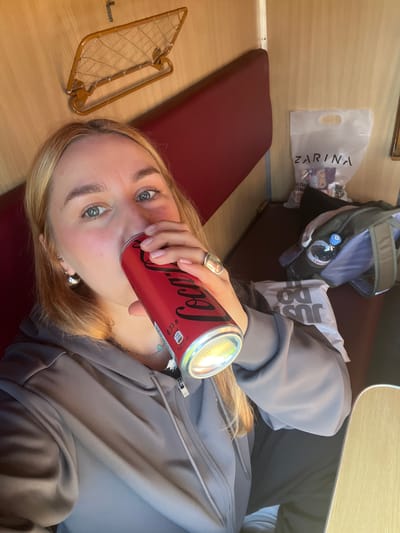 A young woman is drinking a Coca-Cola can while lying down in what appears to be a train compartment. She wears a gray zip-up jacket and has blonde hair. Her right hand, adorned with a silver ring, holds the can near her mouth. Other items visible include a luggage rack with a brown wire grid, a red cushioned seat, a bag labeled "Zarina", and a gray backpack with a water bottle in its side pocket. The location is inferred to be a train traveling through or near Заслаўе, Belarus, based on visual context suggesting a train interior.