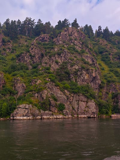 The image depicts a rocky mountainside covered in lush green vegetation and scattered pine trees, sloping down to a wide, dark river. The sky above is overcast with soft, diffused light. The rocks are rugged and uneven, with visible strata and cracks, suggesting a natural geological formation. The vegetation is dense, with various shades of green indicating different types of plants and shrubs clinging to the rocky terrain. The river in the foreground is calm, with subtle ripples on its surface, reflecting the muted colors of the sky and the surrounding landscape. The scene appears to be a natural landscape, likely a riverbank and its adjacent cliff face, located in or near the city of Atamanovo, Russia, given the context of its origin. There are no people or specific man-made objects visible, creating a serene and untouched natural environment.