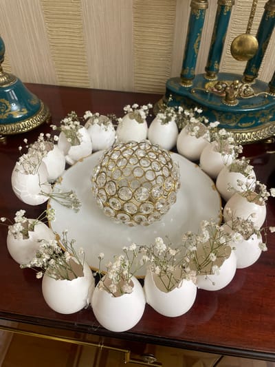 A high-angle, slightly tilted indoor shot depicts a decorative arrangement on a dark brown wooden surface.
The arrangement features a white plate with a gold rim, centered on which is a golden, faceted orb. Surrounding the plate are white eggshells, some cracked open, each containing sprigs of small white flowers.
To the left of the arrangement, the edges of a teal and gold decorative object are visible, partially obscured by the eggshells and plate. The background features a textured cream-colored wall.
The image was taken in the city of Akhalkalaki, Georgia.