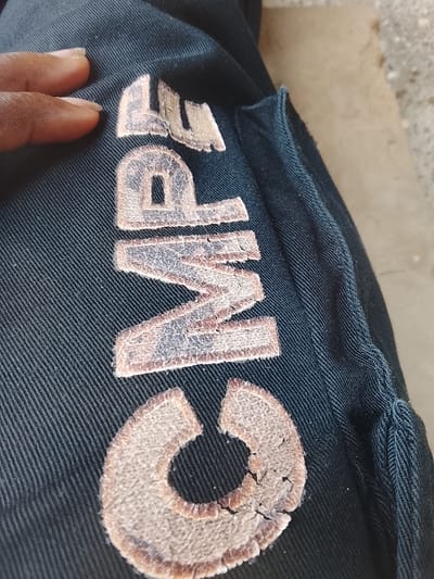 A close-up, high-angle image depicts dark blue woven fabric, possibly a garment, featuring four light beige or tan embroidered letters that spell "CMPE". The letters exhibit a distressed texture with visible cracks and wear. A hand with dark skin, specifically a thumb and part of an index finger, rests on the upper-left portion of the fabric, adjacent to the letter "C." The background consists of an out-of-focus light gray or tan surface, consistent with a concrete ground. The scene is located in Rumuekini, Nigeria.
