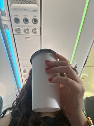 The image appears to be taken inside an airplane. A person is holding a white cup with a dark gray lid. The person has red nail polish, a silver-colored ring, and a gold-colored watch. The person has brown curly hair. The interior of the airplane cabin is visible, including an overhead panel with controls for lights, air, and Bluetooth. There is a blue light strip above the panel. The location in Apodaca, Mexico is not evident from the image.