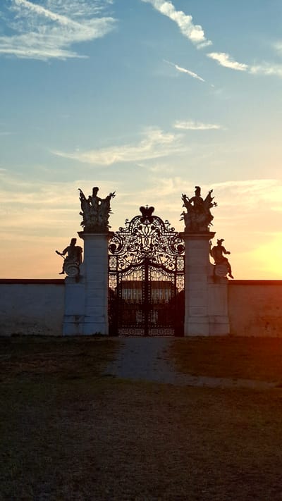 The image depicts the ornate wrought-iron gate of Schloss Hof in Austria at sunset. Silhouetted against a pale blue and orange sky, the gate features intricate scrollwork and is flanked by two white pillars. Each pillar is topped with a statue of a figure, and smaller statues are positioned on the sides of the pillars. The gate opens onto a path that leads to a building, also silhouetted. The foreground consists of a grassy area. The overall scene conveys a sense of historical grandeur and is bathed in the warm light of the setting sun.