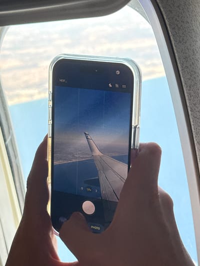A person is holding a smartphone horizontally to take a picture of an airplane wing outside an airplane window. The airplane wing is white and extends from the right side of the phone's screen towards the left. Below the wing, clouds and the earth's surface are visible, appearing as a mixture of brown and white textures, suggesting land and possibly more clouds. The sky is a clear blue. The phone is in a clear protective case. Visible on the phone screen are camera interface elements: zoom levels "0.5", "1x", "19x", and "2", a circular shutter button, and the word "PHOTO". On the bottom left of the screen, "HEIF" is visible, indicating the file format. The image appears to be taken during the daytime, with bright sunlight illuminating the scene. The overall mood is one of capturing a moment of travel. The image was taken in Pentakomo, Cyprus.