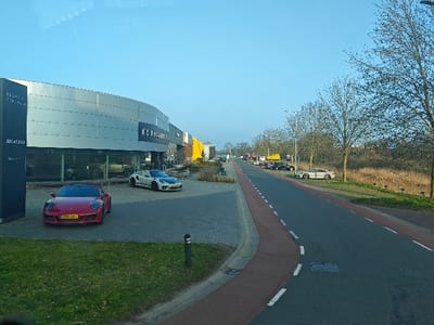 The image displays an exterior scene of the Klaasen & ter Beek B.V. car dealership at Parallelweg 110 in Wierden, Netherlands, under clear daylight. In the foreground, a red Porsche 911 convertible, bearing Dutch license plate HFN-33-J, is parked on a dark gray paved surface. Adjacent to it, a tall, dark vertical sign identifies "KLAASEN & TER BEEK" and "All Car Lease." The dealership building itself is a modern, multi-story structure with a curved facade composed of gray and white panels and large glass display windows revealing multiple luxury vehicles inside. Parked on the paved area directly in front of the building is a white Porsche 911 GT3, featuring black and blue accent graphics.
To the right, a two-lane asphalt road with dashed white center markings and visible manhole covers extends into the distance. A reddish-brown paved bicycle path runs parallel to the road on the left. A row of bare deciduous trees lines the right side of the road, beyond which lies an area of dry, light-brown vegetation. Further down the road, a light-colored sports car with a large rear wing is parked on the right shoulder. Several other vehicles, including cars and trucks, are visible parked along the road in the background. The sky is clear and bright blue. No individuals, actions, or interactions are discernible within the frame.