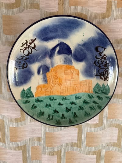 A ceramic plate is depicted, with a dark blue rim. The plate features a painted scene of a building with multiple domes, rendered in orange and white. Green foliage and stylized green flora are also present. Dark, stylized script adorns the upper left and right portions of the plate. The plate rests on a patterned tablecloth, which has alternating bands of light pink, beige, and grey. The image does not contain any information about the city of Golenishchevo, Russia.