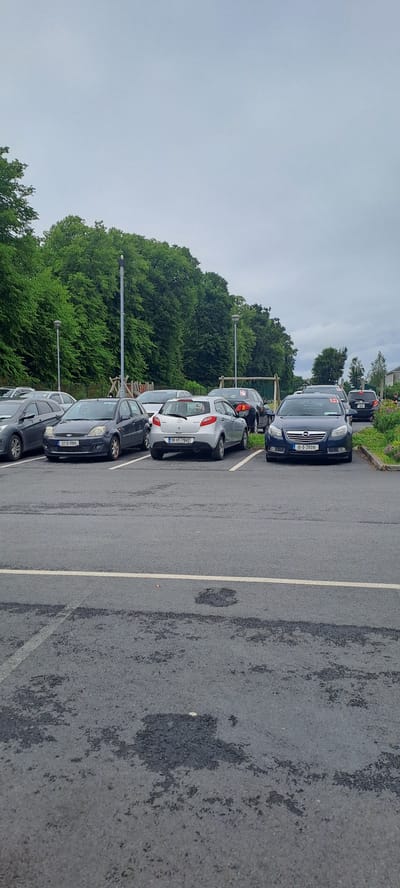 The image shows a parking lot in Breaffy, Ireland, with several cars parked in designated spaces. The lot is paved with asphalt and marked with white lines. In the background, there is a dense line of trees, suggesting a rural or park-like setting. Several streetlights are visible, indicating the area is illuminated at night. Some of the cars have red stickers on their windshields, possibly for an event or marking. The sky is overcast, and the overall lighting suggests it is daytime.