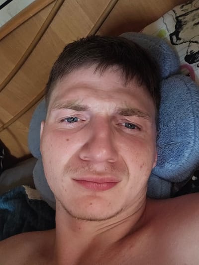 The media file is a close-up, rotated selfie of a person lying down. The subject appears to be a young adult with light skin and short, dark hair. They have blue eyes, light eyebrows, and some stubble on their chin and jawline. Their mouth is slightly open, revealing pink lips. The person is resting their head on a blue, fluffy pillow, which is placed on a patterned surface. In the background, there are wooden planks, suggesting an indoor setting, possibly a bed or furniture. The lighting is soft and diffused, indicating it could be daytime or a well-lit room. The overall mood seems relaxed, perhaps captured in a moment of rest or waking up. The location is Dūkštas, Lithuania, though no specific landmarks are visible. There is no visible text in the media file.