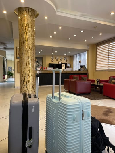 This media file depicts the interior of a hotel lobby. The scene is oriented vertically, showing a perspective looking upwards from the floor. In the foreground, two suitcases are positioned upright, with the larger, light blue suitcase in the back and a gray suitcase in front. The blue suitcase has "Reebok" printed on it. A black backpack is also visible behind the suitcases.

The lobby itself features a reception desk in the background with what appears to be a set of world clocks mounted on the wall behind it. Seating areas with red chairs and sofas are visible to the left of the reception desk. The walls are adorned with various lights, and a large, textured pillar with a mosaic-like pattern stands prominently in the middle ground. Framed artwork and a potted plant can also be seen in the lobby.

The lighting suggests it is daytime, and the indoor setting eliminates any information about weather conditions. The overall atmosphere appears calm and unoccupied, suggesting a quiet moment in the hotel lobby, possibly during check-in or check-out. The location cues, such as the world clocks and the general decor, are consistent with a hotel setting. No people are actively visible in the frame.